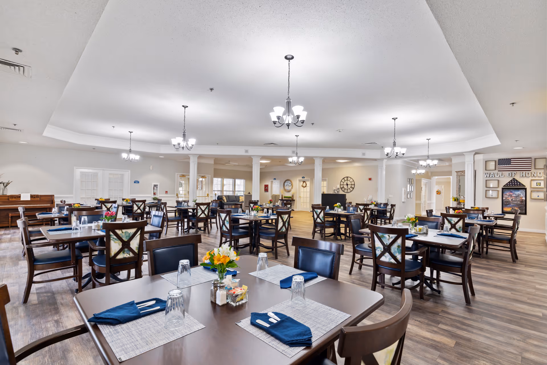 A spacious dining room with multiple dark wood tables and chairs arranged neatly. Each table is set with placemats, blue napkins, utensils, and upside-down glasses. The room features wood flooring, white walls, and several ceiling light fixtures. In the background, there is a piano on the left side and a 'Wall of Honor' display with an American flag on the right side.