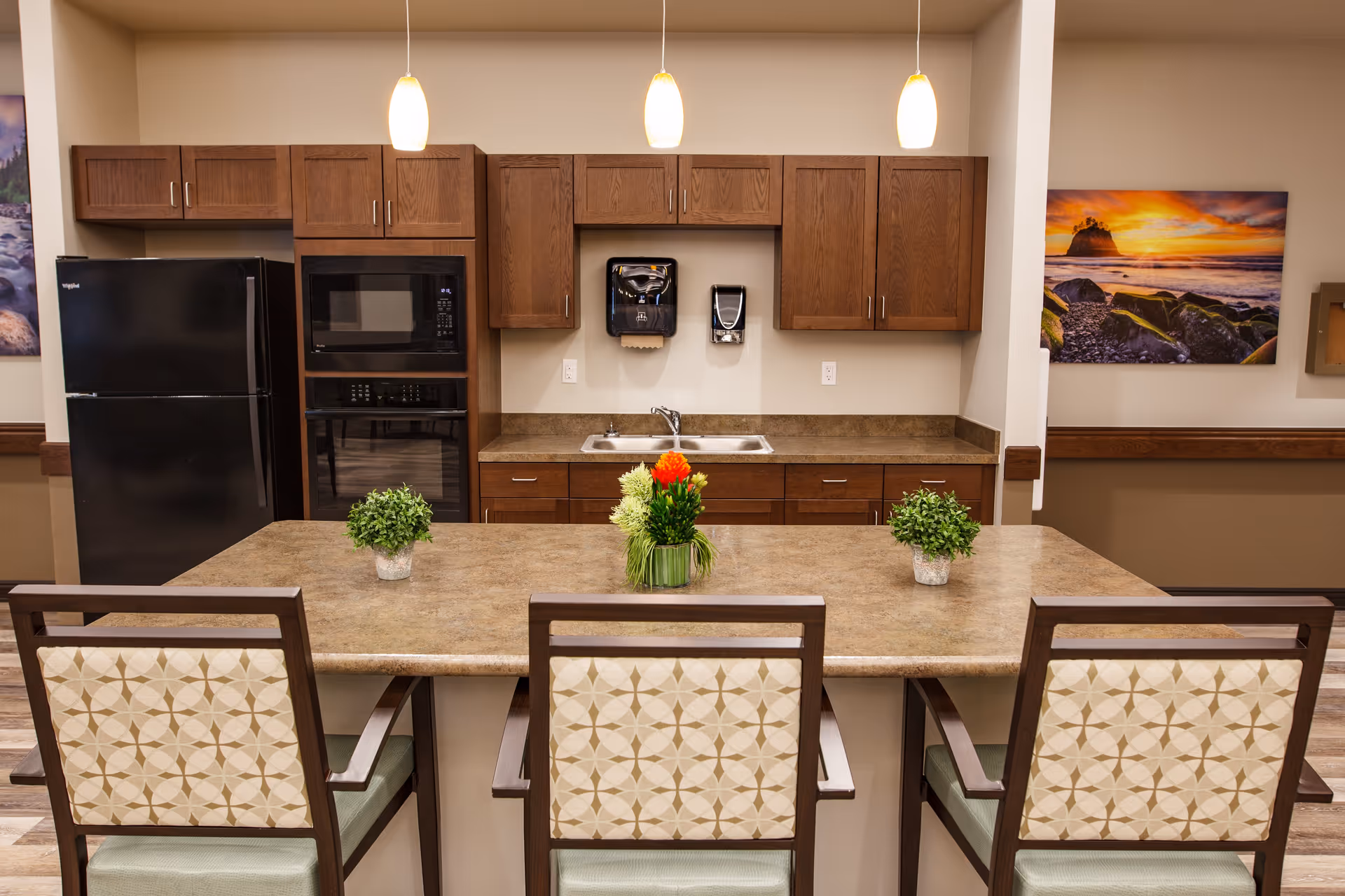 A kitchen area in a senior living facility with a countertop island featuring three chairs with patterned backs. The kitchen has wooden cabinets, a black refrigerator, a built-in microwave and oven, a double sink, and two wall-mounted dispensers. Three pendant lights hang above the island, and there are decorative plants on the countertop. A colorful landscape painting is visible on the wall to the right.
