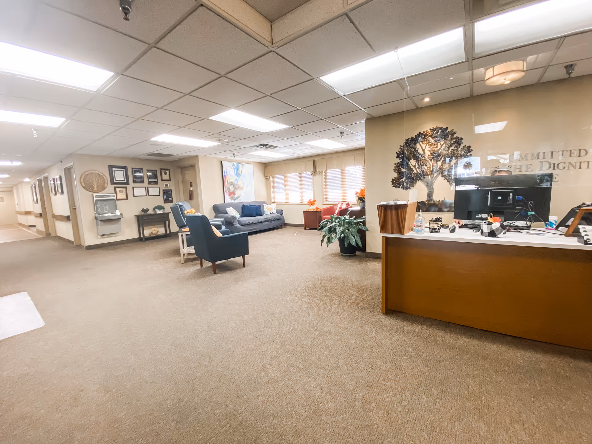 Interior view of a senior living facility lobby or common area with a reception desk on the right, a seating area with chairs and a sofa in the center, and a hallway leading to rooms on the left. The walls are decorated with framed pictures and a large tree artwork behind the reception desk. The area is well-lit with ceiling lights and has carpeted flooring.