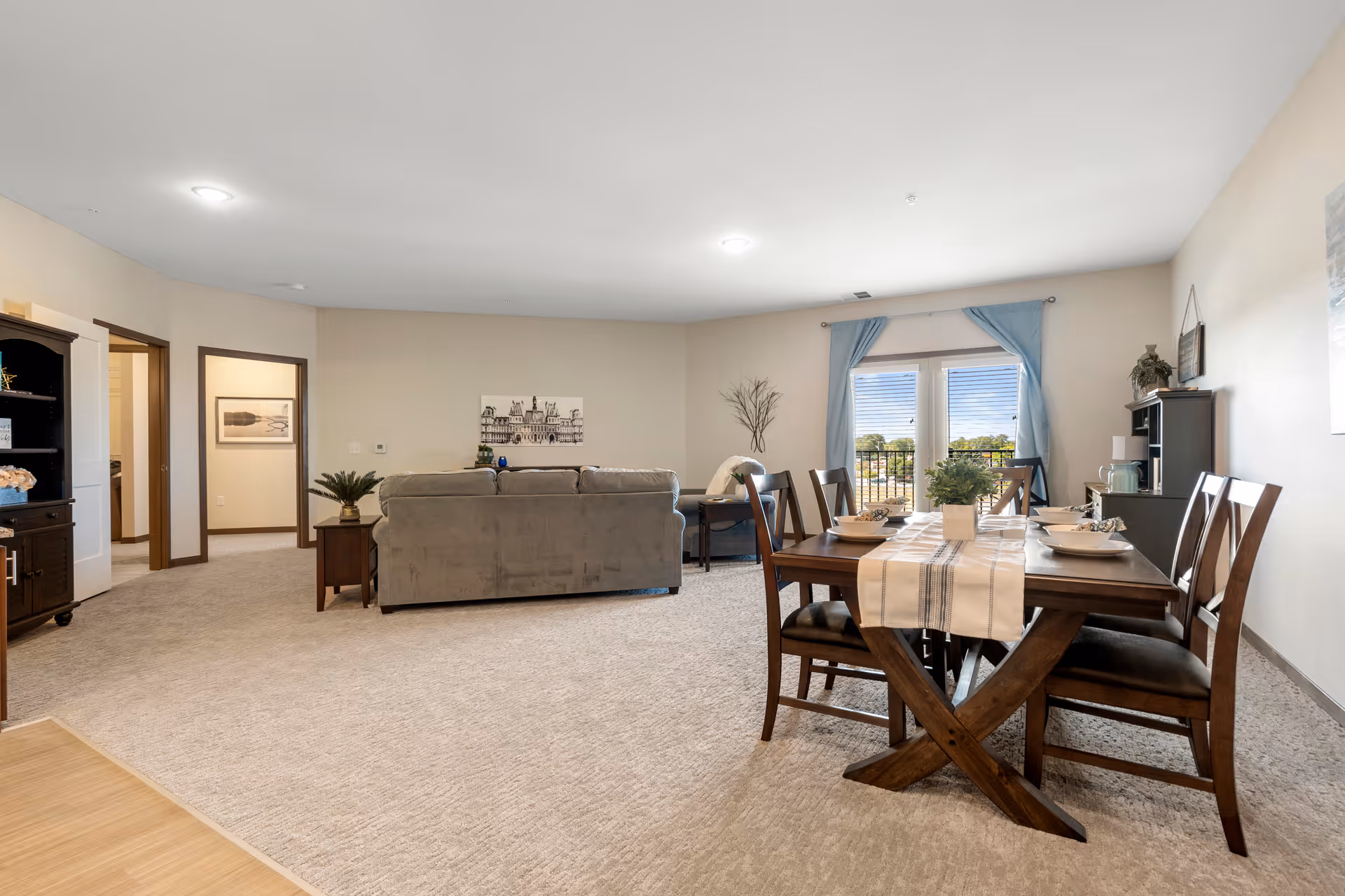 A spacious living and dining area in a senior living facility with beige walls and carpeted floors. The dining area features a wooden table with six chairs, set with plates and a table runner. The living area has a gray sofa facing away from the camera, a side table with a plant, and a small desk with a chair near a window with blue curtains. The room is well-lit with ceiling lights and natural light from the window.