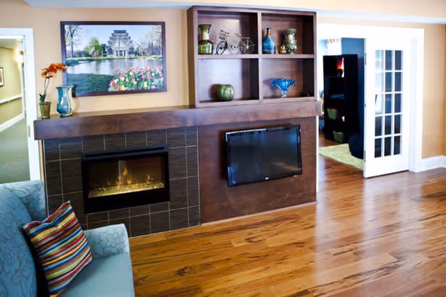 A cozy living room area featuring a modern fireplace with a wooden mantel, a mounted flat-screen TV, and built-in shelves displaying decorative vases and a small bicycle figurine. There is a light blue armchair with a colorful striped pillow on the left side. The room has wooden flooring and beige walls, with a hallway and a room with French doors visible in the background.