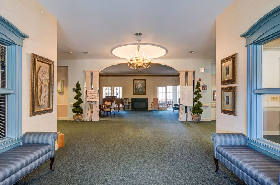 Spacious interior common area with blue cushioned benches on either side, framed artwork on the walls, potted plants, and a chandelier hanging from the ceiling. In the background, there is a seating area with chairs and tables near windows and a fireplace.