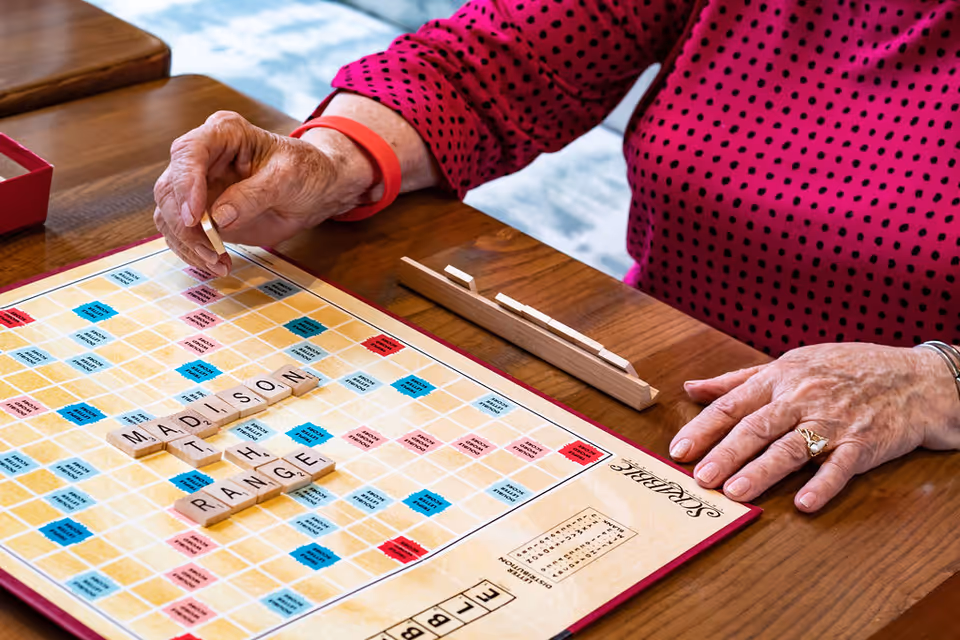 An elderly person wearing a red polka dot shirt is playing Scrabble at a wooden table. The Scrabble board has the words 'MADISON AT THE RANGE' spelled out. The person's hands are visible, one holding a Scrabble tile and the other resting on the table near the tile rack.