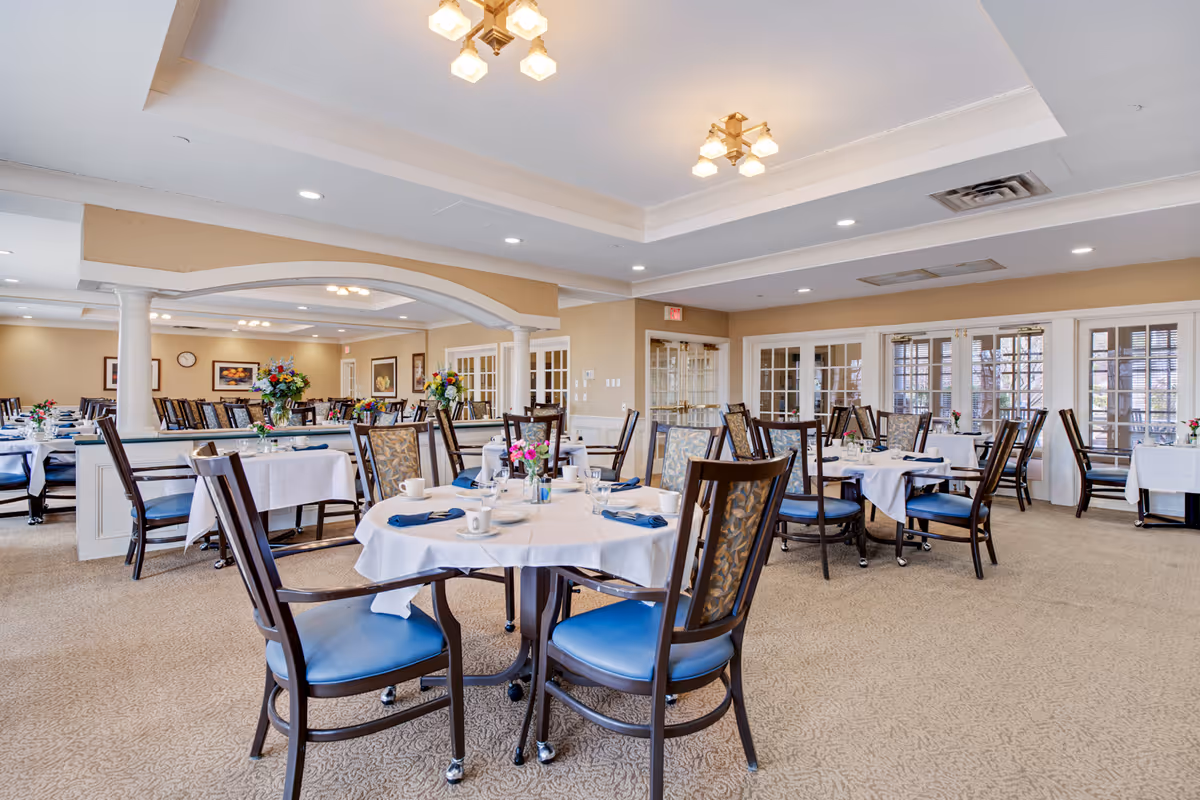 A spacious dining room in a senior living facility with multiple round tables covered with white tablecloths. Each table is set with cups, plates, and blue napkins. The room features beige walls, carpeted floors, and large windows with glass doors allowing natural light to enter. There are floral centerpieces on the tables and elegant ceiling light fixtures.