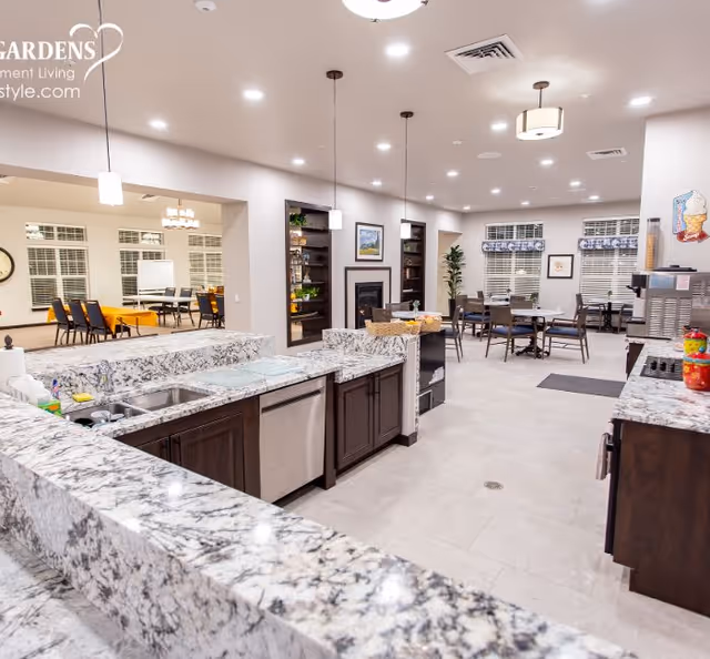 A spacious and well-lit common area in a retirement living facility featuring a granite countertop kitchen with stainless steel appliances in the foreground. The room includes multiple dining tables and chairs, a fireplace with built-in shelves, and large windows with blinds. Pendant lights hang from the ceiling, and there is an adjacent room visible with additional seating and a clock on the wall.
