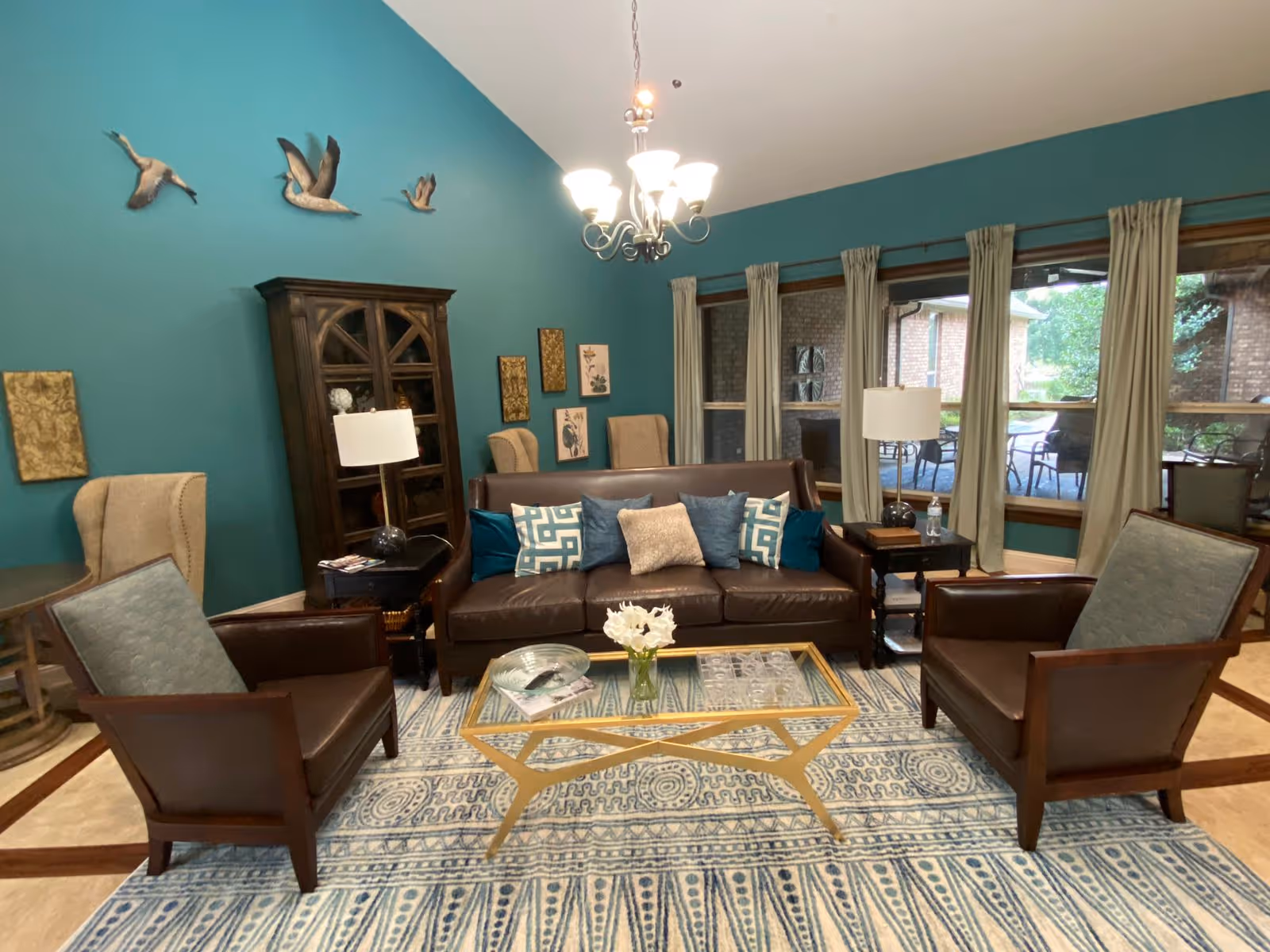 A cozy living room with teal walls featuring decorative bird sculptures and framed artwork. The room has a brown leather sofa with multiple blue and beige cushions, two brown armchairs with light blue upholstery, a glass coffee table with a gold frame, and a patterned blue and white rug. There are two side tables with lamps and a wooden cabinet against the wall. Large windows with beige curtains provide a view of an outdoor patio area with chairs and greenery.