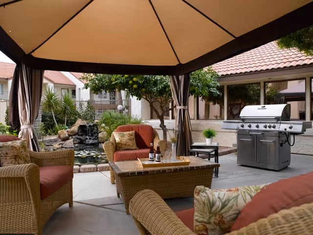 Outdoor patio area with wicker furniture including chairs and a coffee table under a large canopy. There is a stainless steel barbecue grill on the right side and a small pond with a waterfall feature surrounded by greenery in the background. The setting is part of a residential facility with buildings visible in the background.