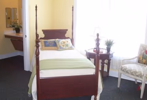 A small bedroom with a wooden four-poster single bed made with white and green bedding and decorative pillows. Next to the bed is a wooden side table with a lamp and a small plant. A patterned upholstered chair is positioned near a window with sheer white curtains. In the background, a sink and mirror are visible in an adjoining bathroom area.