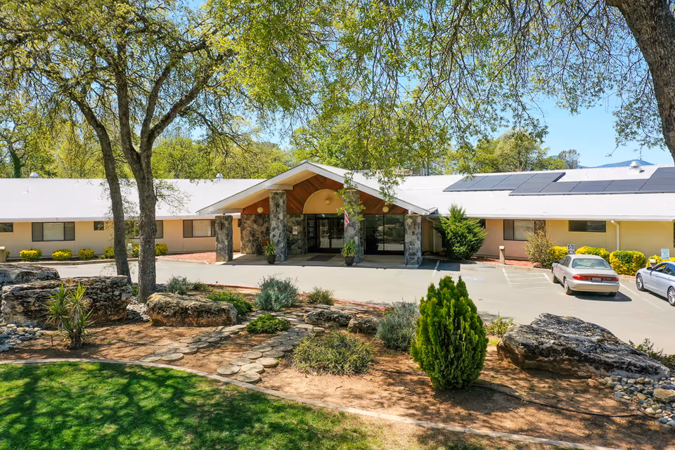 Single-story building front with a covered entrance supported by stone columns, a landscaped rock garden in the foreground, and parked cars.