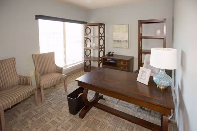 Bright furnished office-style room with a wooden desk, two striped chairs by a large window, shelving units, and a table lamp.