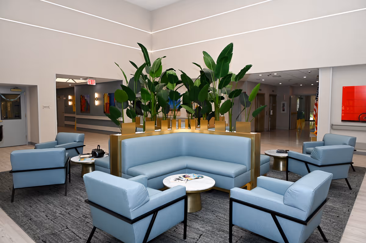 A modern seating area in a senior living facility featuring a light blue sectional sofa and matching armchairs arranged around small round tables. Tall green plants in gold planters are placed in the center of the sectional, creating a natural divider. The space has a neutral color palette with light walls and a gray carpet, and there are hallways and artwork visible in the background.