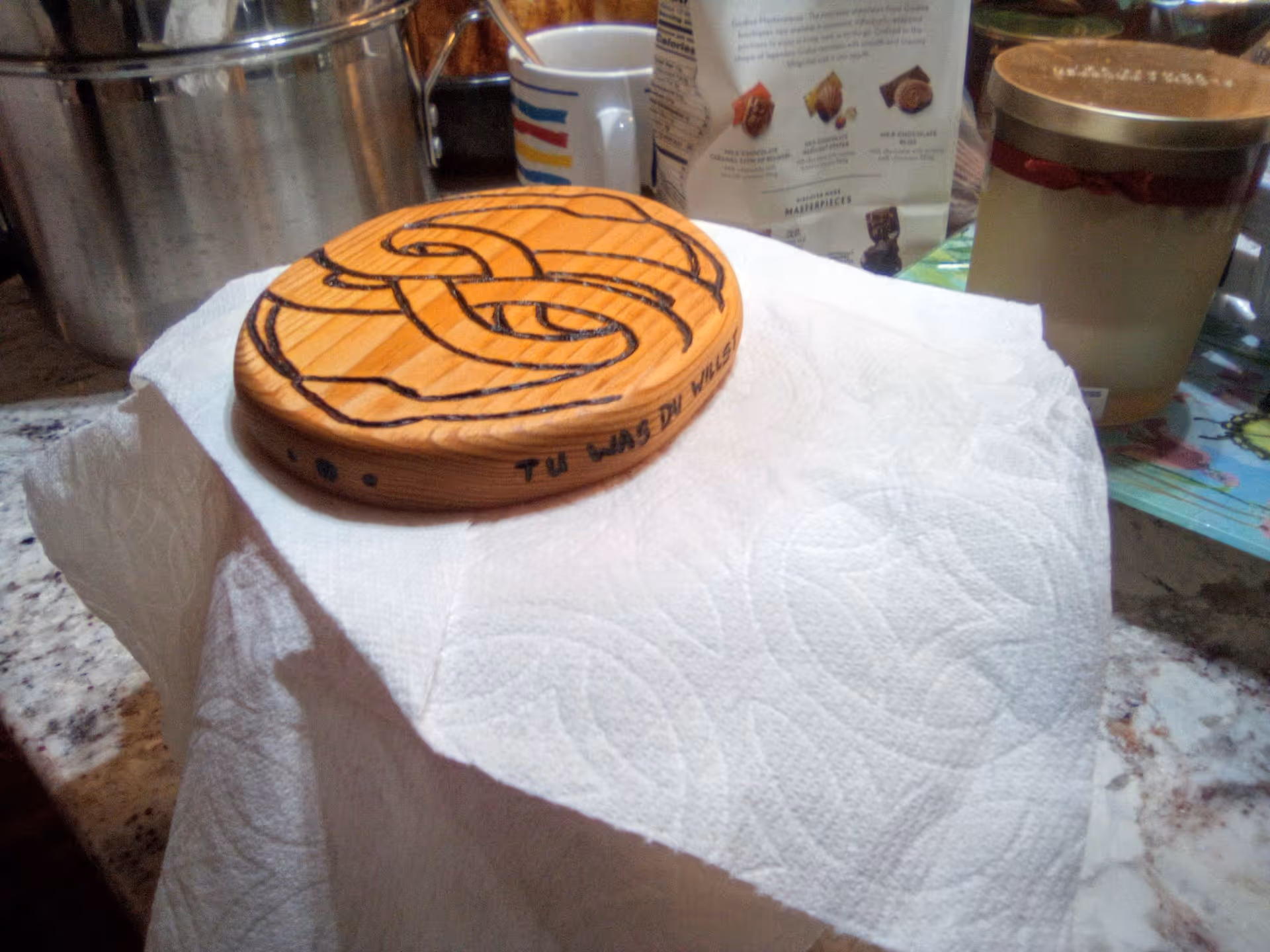 A carved wooden coaster with a knot design sits on a paper towel on a kitchen countertop with pots and jars in the background.