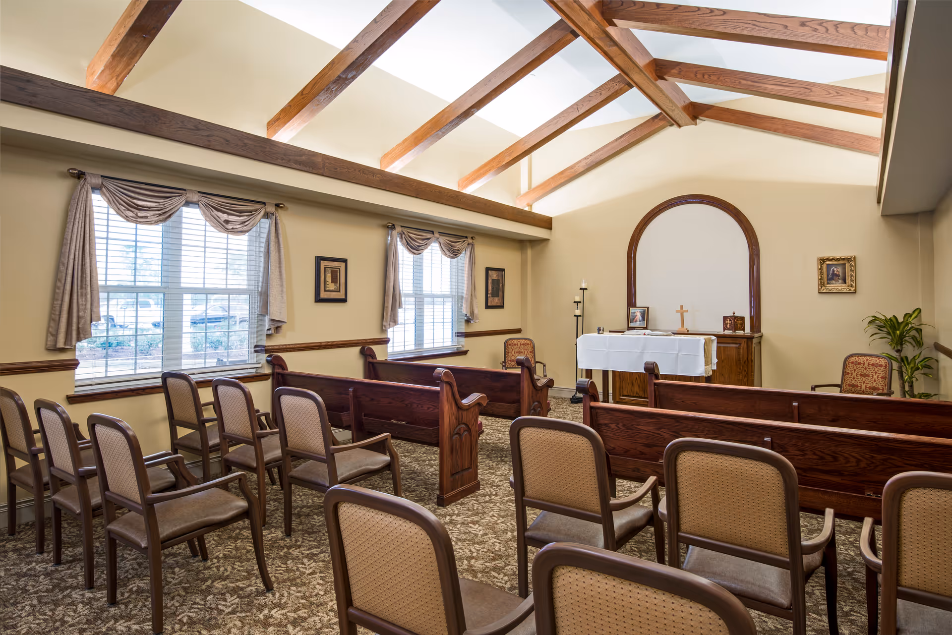 Interior of a small chapel or prayer room with wooden pews and chairs arranged facing a small altar covered with a white cloth. The room has large windows with curtains, exposed wooden ceiling beams, and framed pictures on the walls.