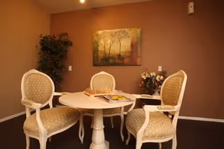 A cozy interior room with a round white table surrounded by three upholstered chairs with patterned fabric. On the table, there is a chessboard and some magazines. The walls are painted brown, and there is a large framed painting of trees hanging on the wall. A potted plant is placed in the corner, and a small table with a flower arrangement is visible in the background.