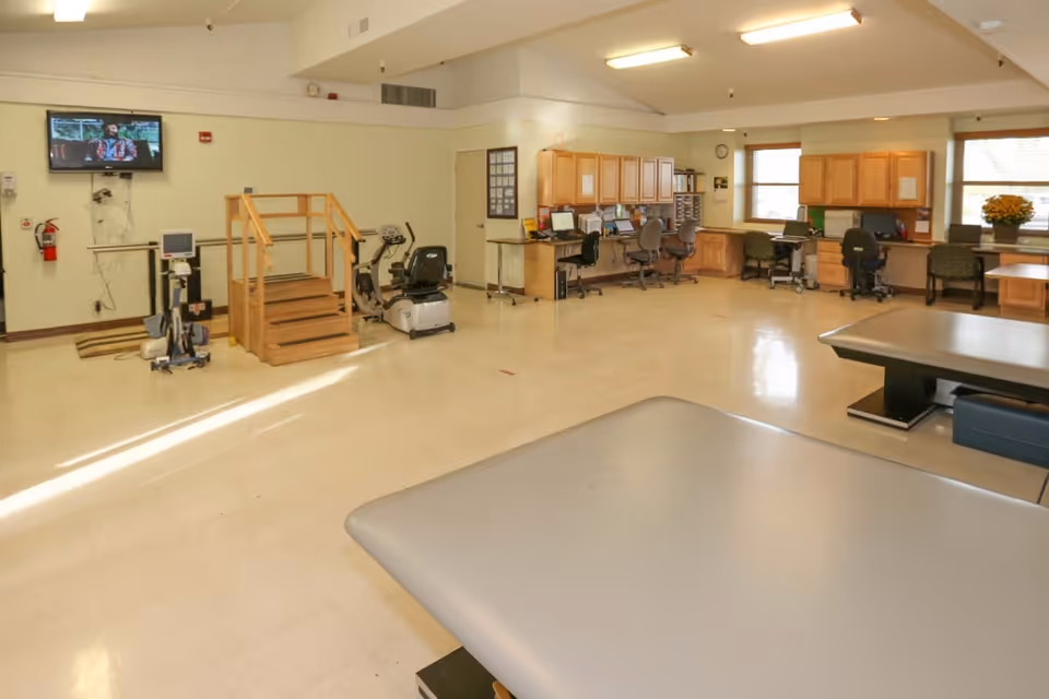 A spacious rehabilitation room with exercise equipment including a stationary bike and a small wooden staircase for physical therapy. There are multiple desks and chairs along the walls with computers and office supplies. The room has large windows letting in natural light and a television mounted on the wall.