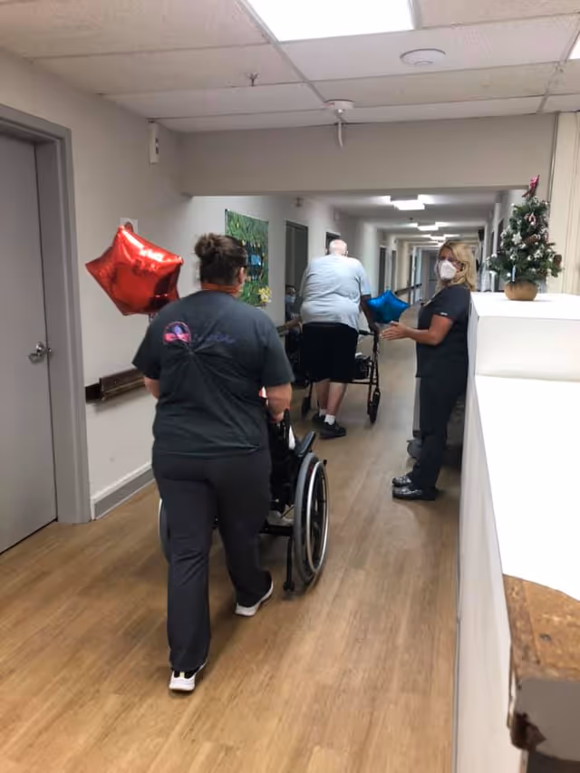 A hallway in a nursing and rehabilitation facility with two healthcare workers assisting elderly patients. One worker is pushing a wheelchair, and another is standing near a small decorated Christmas tree on a counter. The hallway has wooden flooring, white walls, and several doors along the sides.