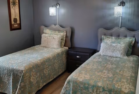 A bedroom with two twin beds dressed in teal patterned bedspreads, a shared nightstand between them, and wall-mounted lamps above each headboard.