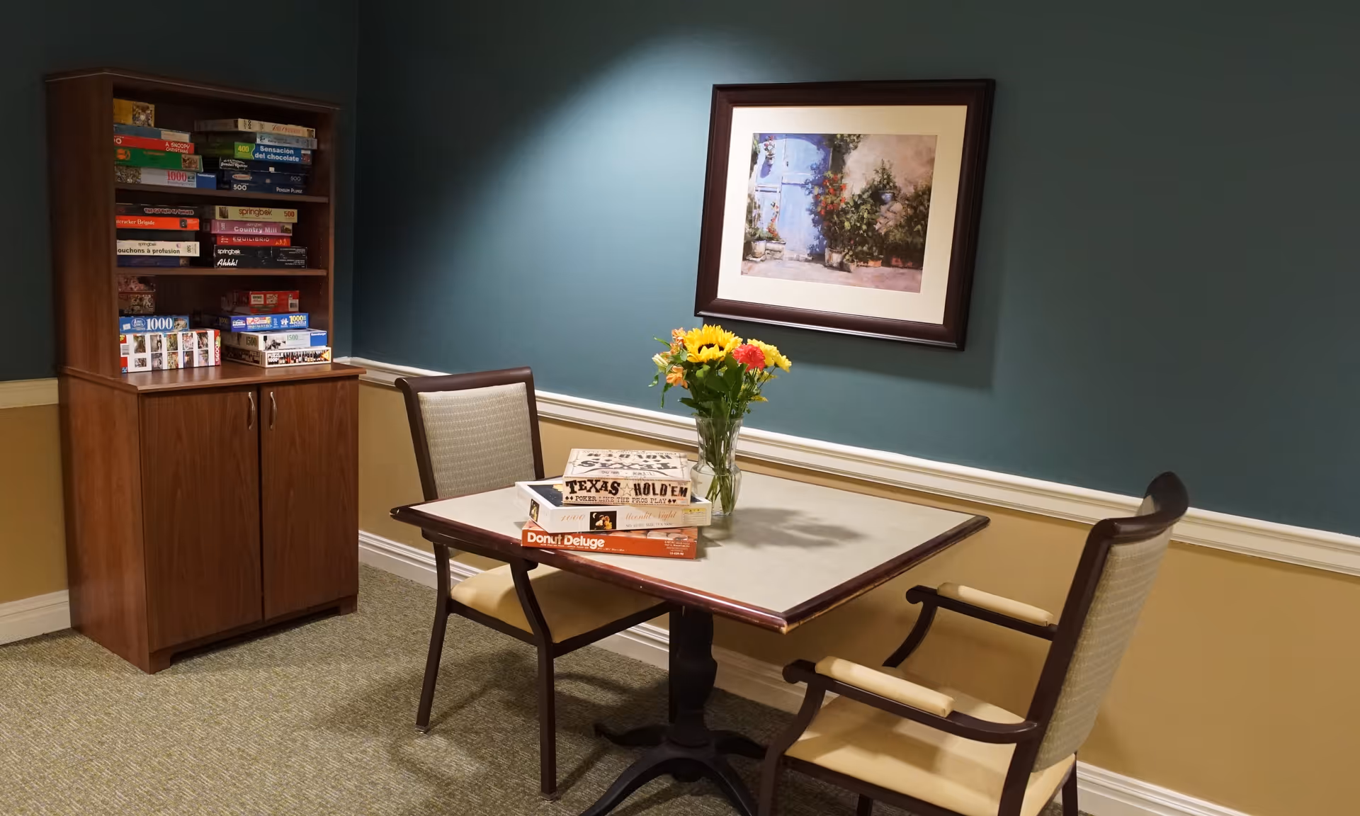 A small seating area with a table and two chairs, a vase of flowers, and a cabinet filled with board games beneath framed artwork on a teal wall.