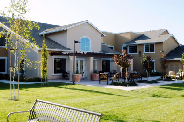 Well-maintained lawn and outdoor patio seating in front of a two-story beige senior living building.