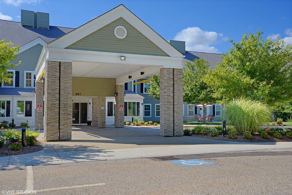 Entrance of a senior living facility with a covered drop-off area supported by stone pillars. The building has light green siding with white trim and multiple windows. There are landscaped areas with bushes, flowers, and trees around the entrance. A handicapped parking space is visible in the foreground under a clear blue sky.