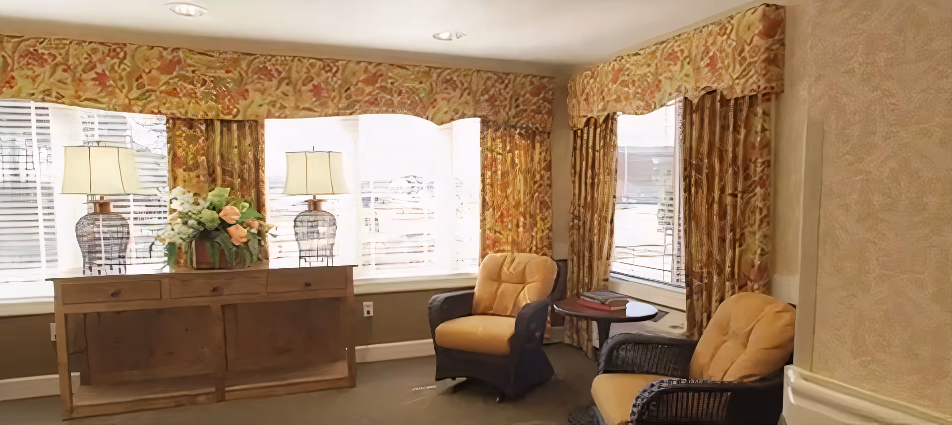 A cozy sitting area with two cushioned armchairs and a small round table between them. Behind the chairs are three large windows with floral-patterned curtains and valances. A wooden console table with two lamps and a flower arrangement is placed in front of the windows. The room has soft lighting and a warm, inviting atmosphere.