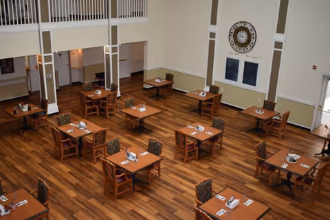 A spacious dining room with multiple wooden tables and chairs arranged neatly on a polished wooden floor. Each table is set with napkins and utensils. The room has high ceilings, light-colored walls, and a large decorative clock on one wall. There is a balcony or upper level visible with white railings.