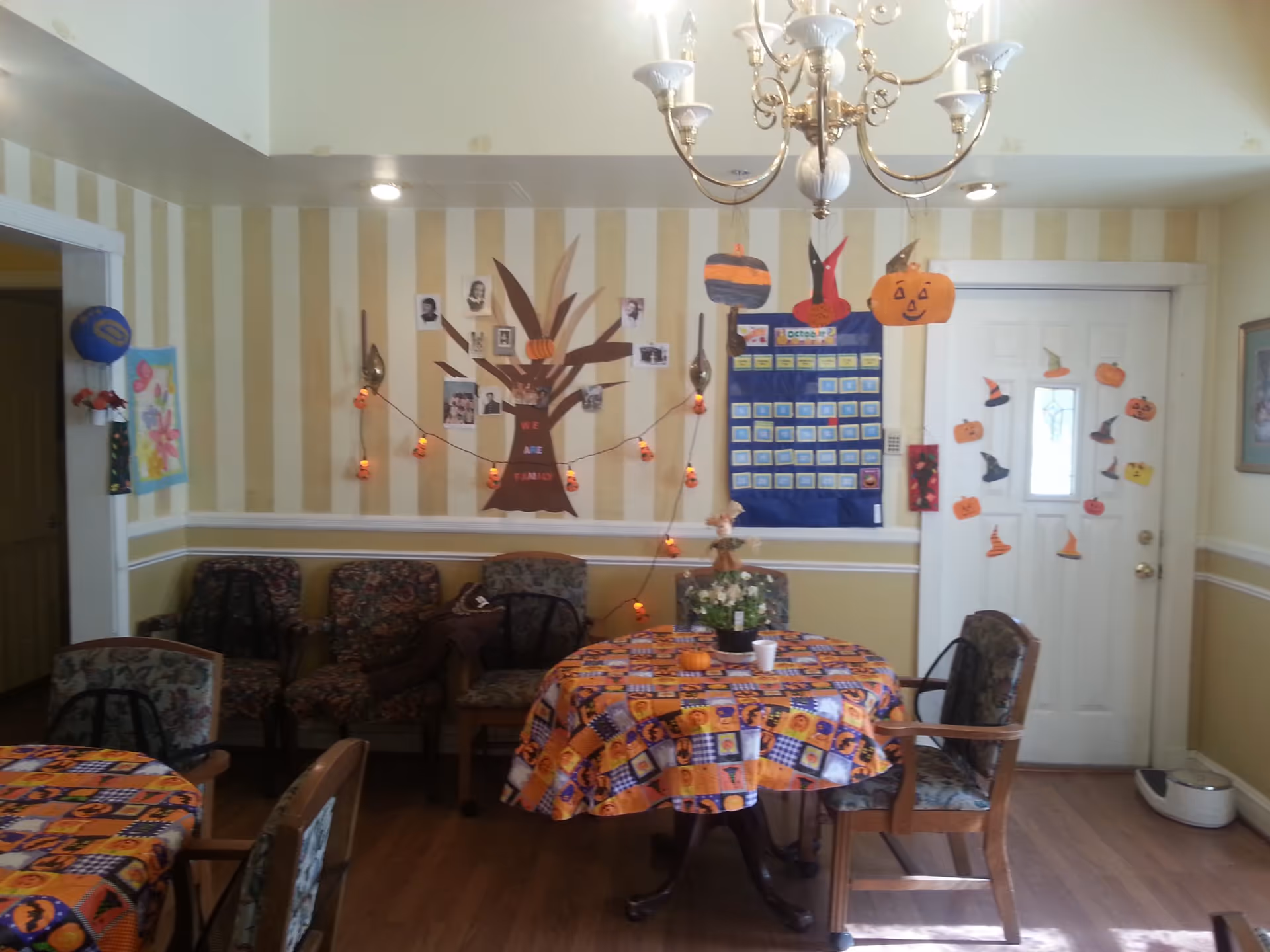 Communal dining/activity room decorated for Halloween with round tables covered in festive tablecloths, chairs, a wall calendar and paper pumpkin decorations.