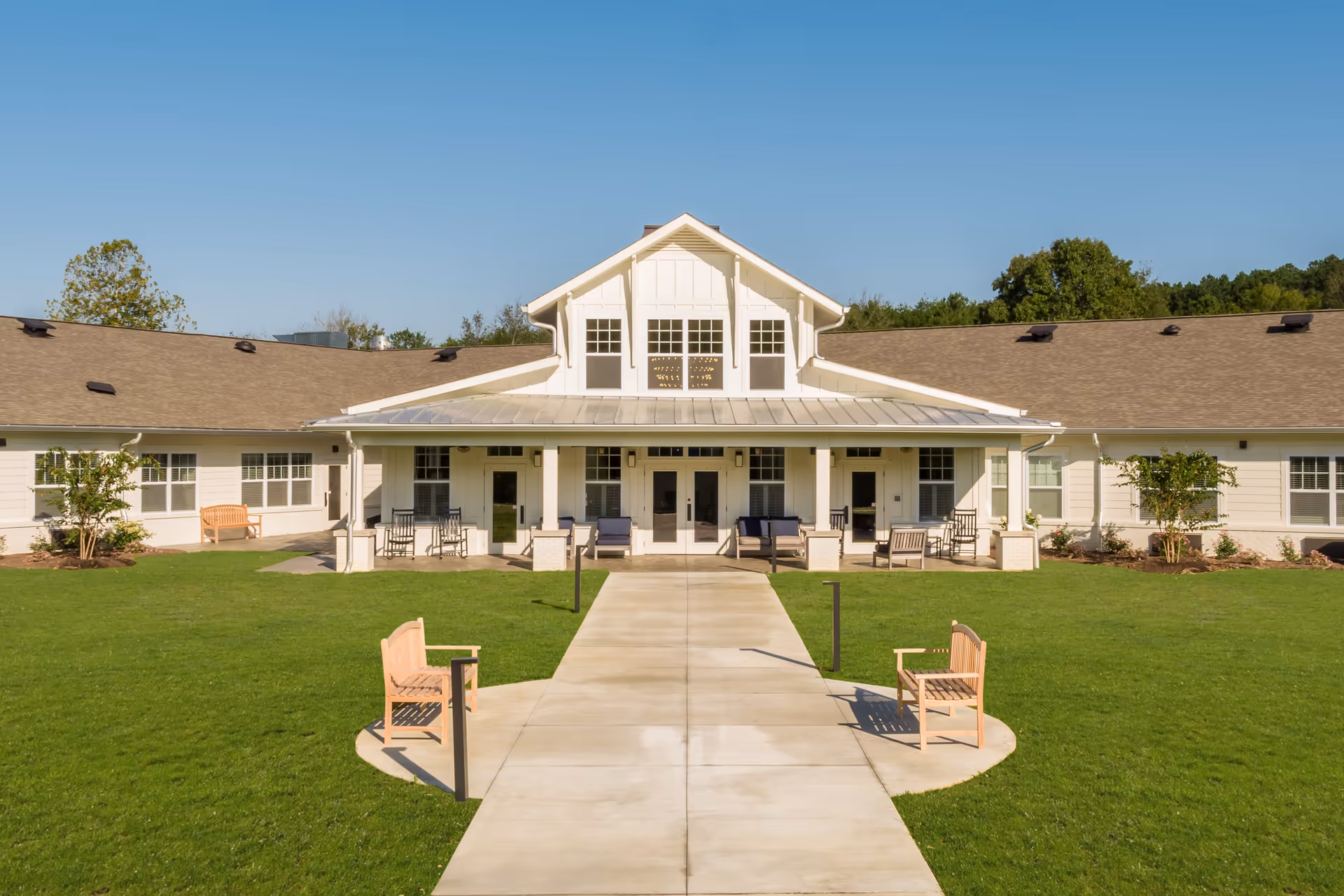 Front exterior view of Dominion Senior Living of Athens building with a wide concrete walkway leading to a covered porch area. The building is single-story with a pitched roof and white siding. There are several wooden benches and chairs placed on the porch and along the walkway. The lawn is green and well-maintained, with a clear blue sky overhead.