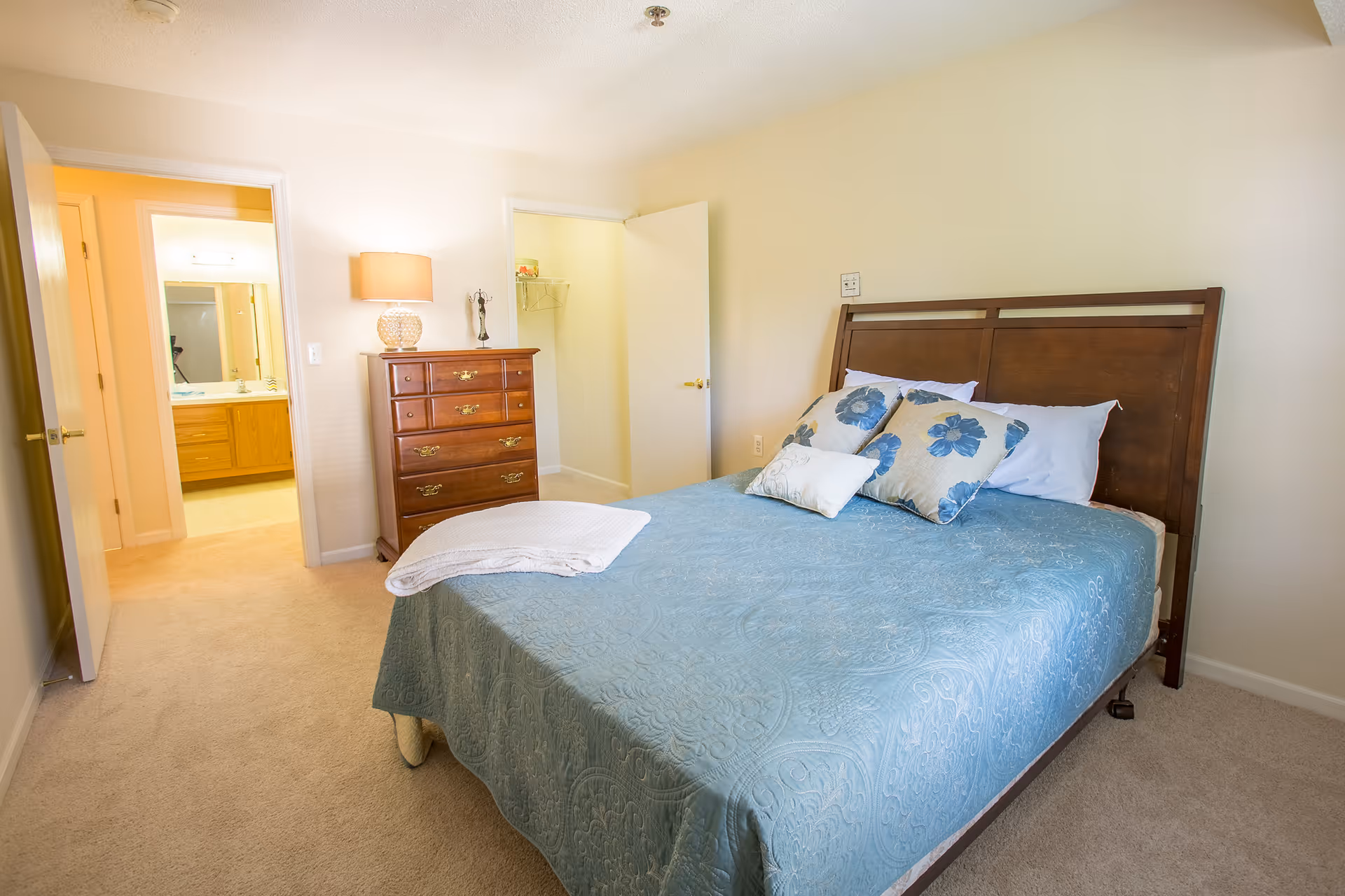 Sunlit bedroom with a made bed in a blue coverlet, wooden headboard, dresser with a lamp, and doors opening to a bathroom and closet.