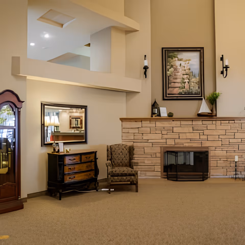 Cozy senior living lounge with a stone fireplace, upholstered armchair, wooden chest, mirror and wall art.