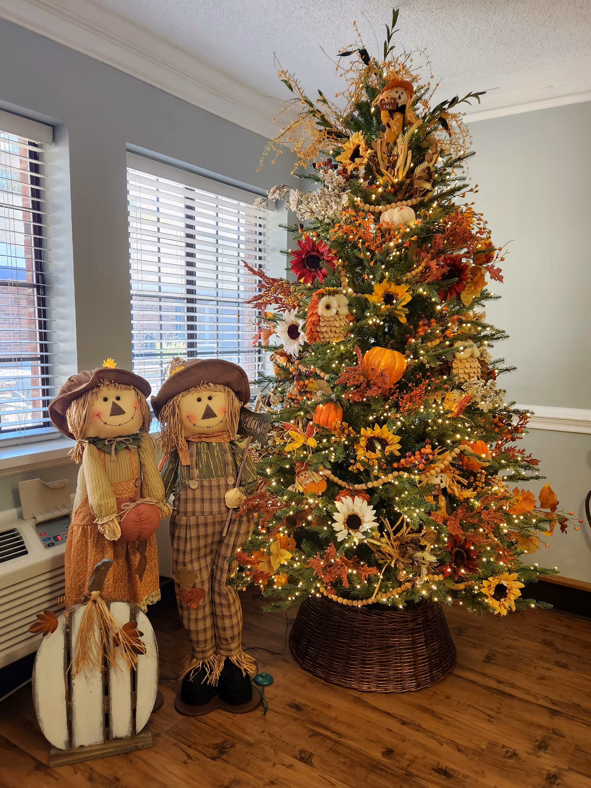 A festive fall-themed decorated Christmas tree with lights, sunflowers, pumpkins, and autumn leaves stands next to two scarecrow figures dressed in autumn attire, one holding a 'Welcome' sign. The scene is set indoors near windows with blinds and a wooden floor.