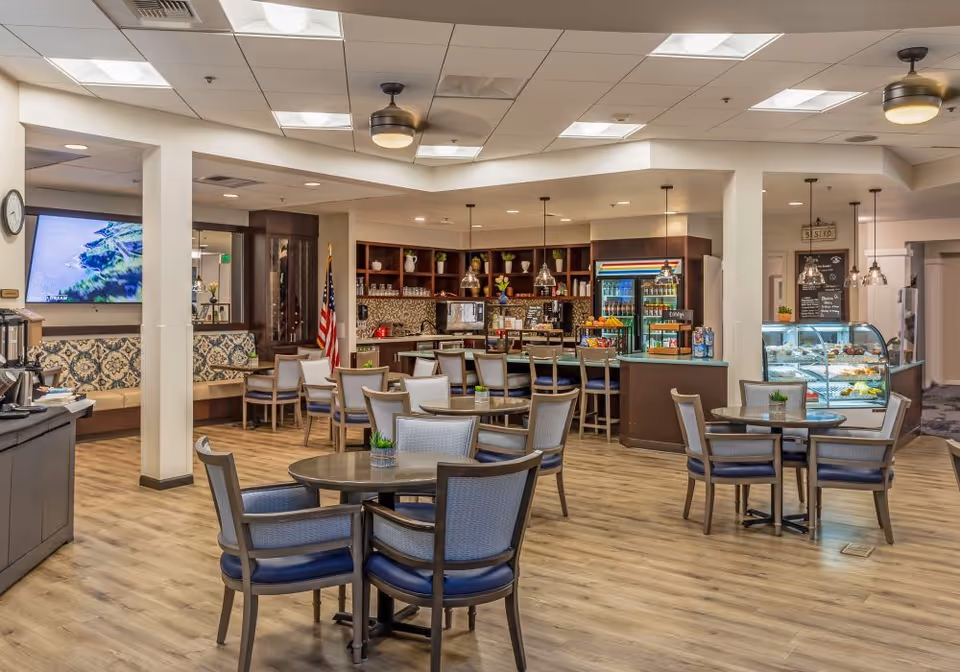 A spacious and well-lit dining area in a senior living facility featuring multiple round tables with chairs, a counter with bar stools, a display case with food items, and a beverage refrigerator. The room has wood flooring, ceiling fans, and a large TV mounted on the wall. The decor includes plants and patterned cushions on a bench seat along the wall.