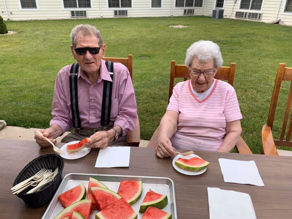 Two elderly individuals sitting outdoors at a table enjoying slices of watermelon. The man on the left is wearing sunglasses, a pink shirt, and suspenders, while the woman on the right is wearing glasses and a pink striped shirt. There is a tray of watermelon slices and a basket of plastic forks on the table. The background shows a grassy courtyard area with windows of a building.