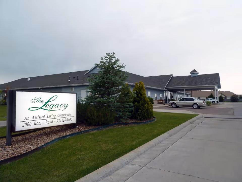 Front exterior of The Legacy assisted living facility showing the entrance, sign, driveway, and parked cars.