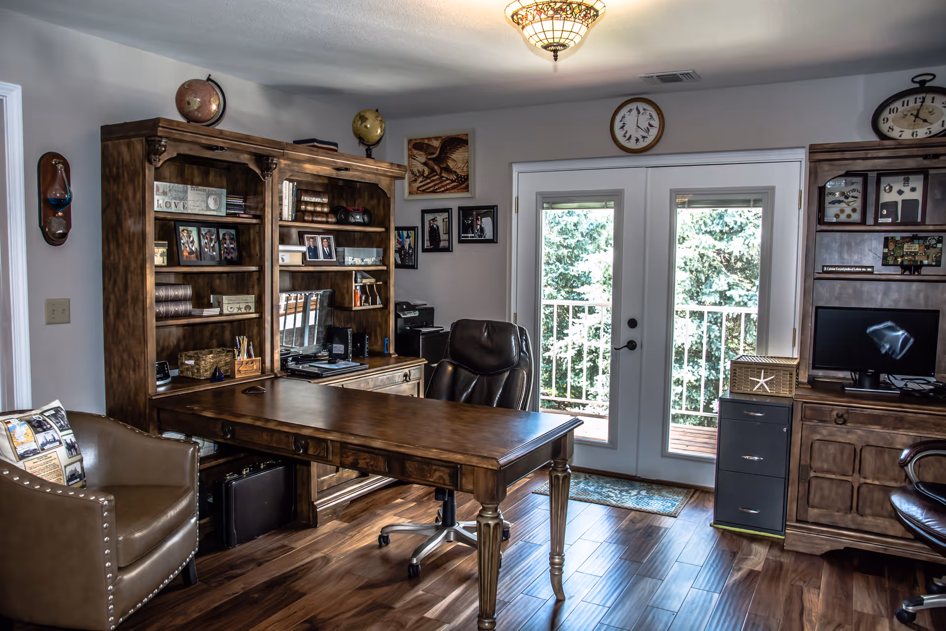 A well-appointed home office with a large wooden desk, bookshelves, leather chairs, and glass double doors opening to a balcony.
