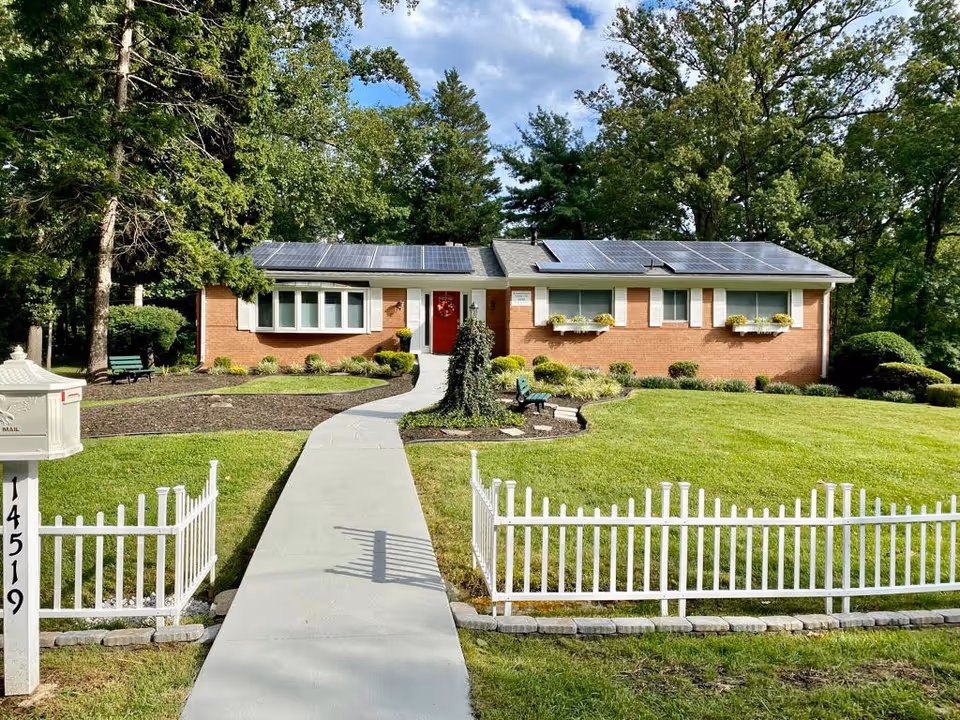 Front exterior view of a single-story brick house with a red door, white window shutters, and solar panels on the roof. The house is surrounded by a well-maintained lawn, flower beds, and trees. A white picket fence and mailbox with the number 14519 are visible in the foreground, along with a concrete walkway leading to the front door.