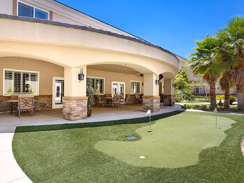 Covered patio with chairs and tables overlooking a small putting green and palm trees at a senior living facility.