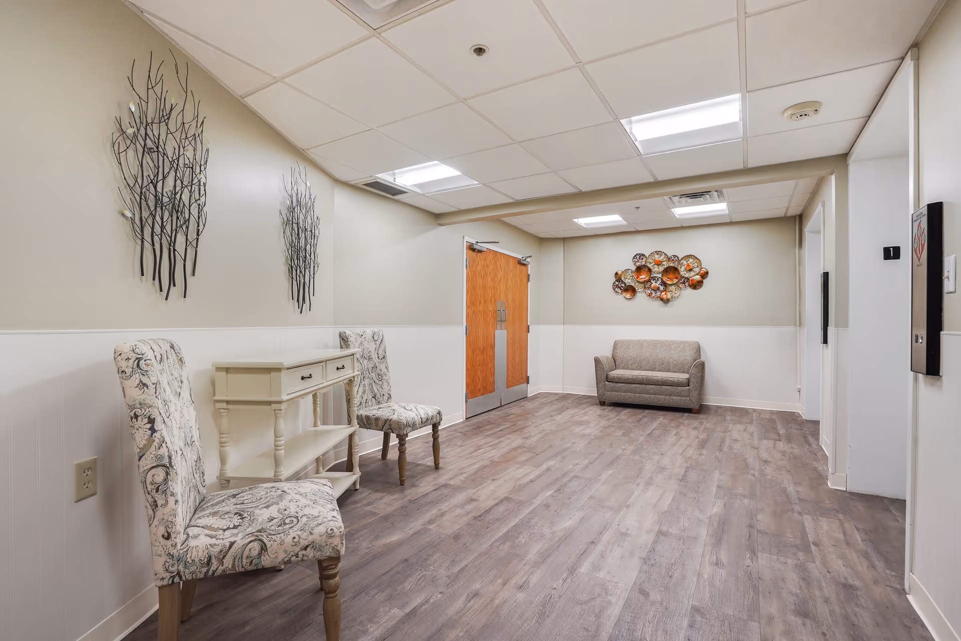 A bright furnished hallway sitting area with two patterned chairs and a small console on the left, a loveseat at the back, and decorative wall art.