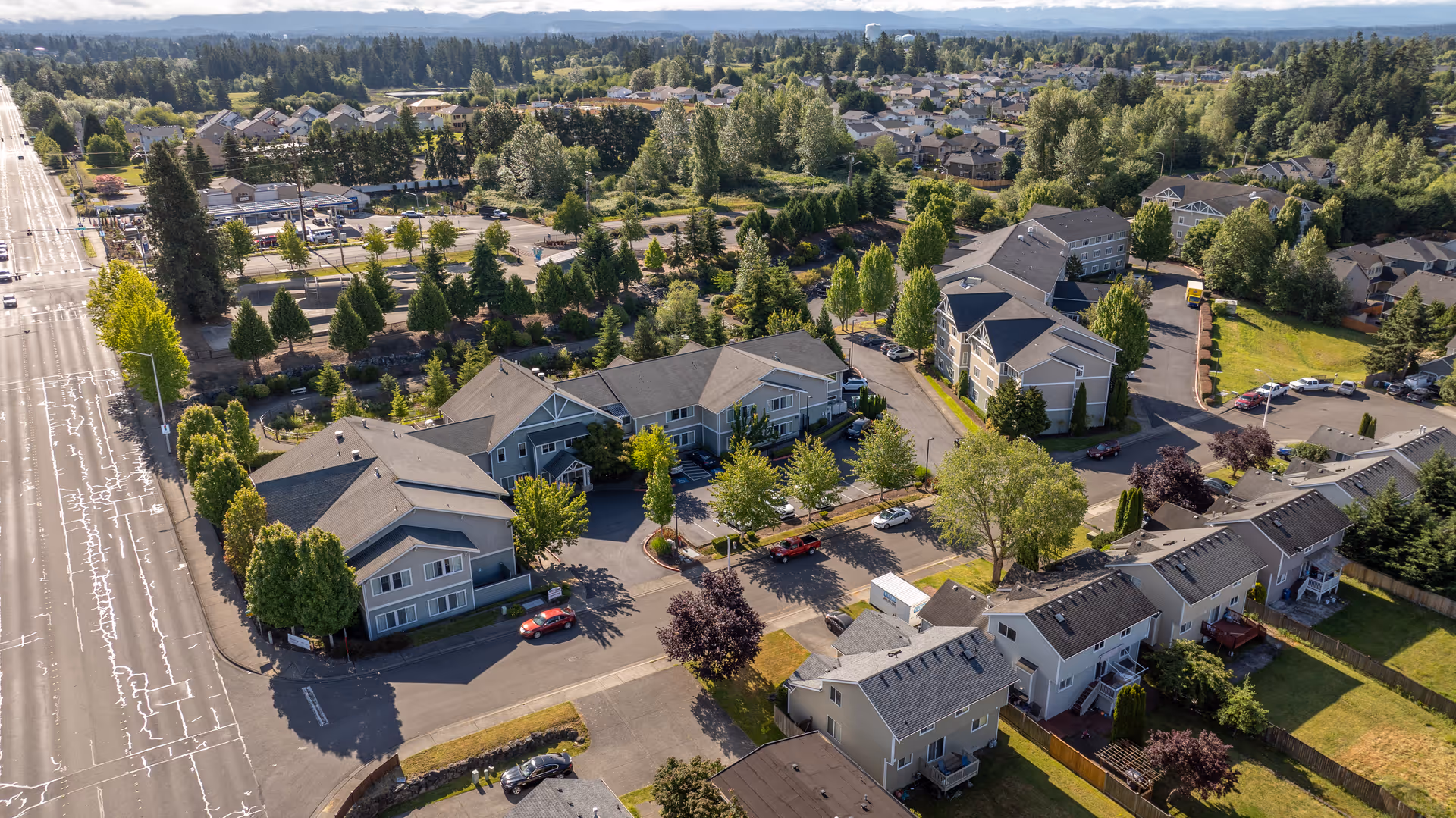 Aerial view of Arbor Village Senior Living facility showing multiple residential buildings surrounded by trees and greenery, with a large road on the left side and a suburban neighborhood in the background under a clear sky.