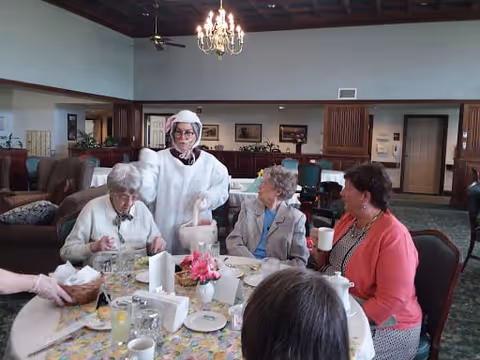 A group of elderly women sitting around a dining table in a communal dining area. One woman dressed in a white costume stands behind the table, while the others are seated and engaged in conversation. The table is set with plates, glasses, and a small flower arrangement. The room has high ceilings with a chandelier and wooden beams, and there are couches and framed pictures in the background.