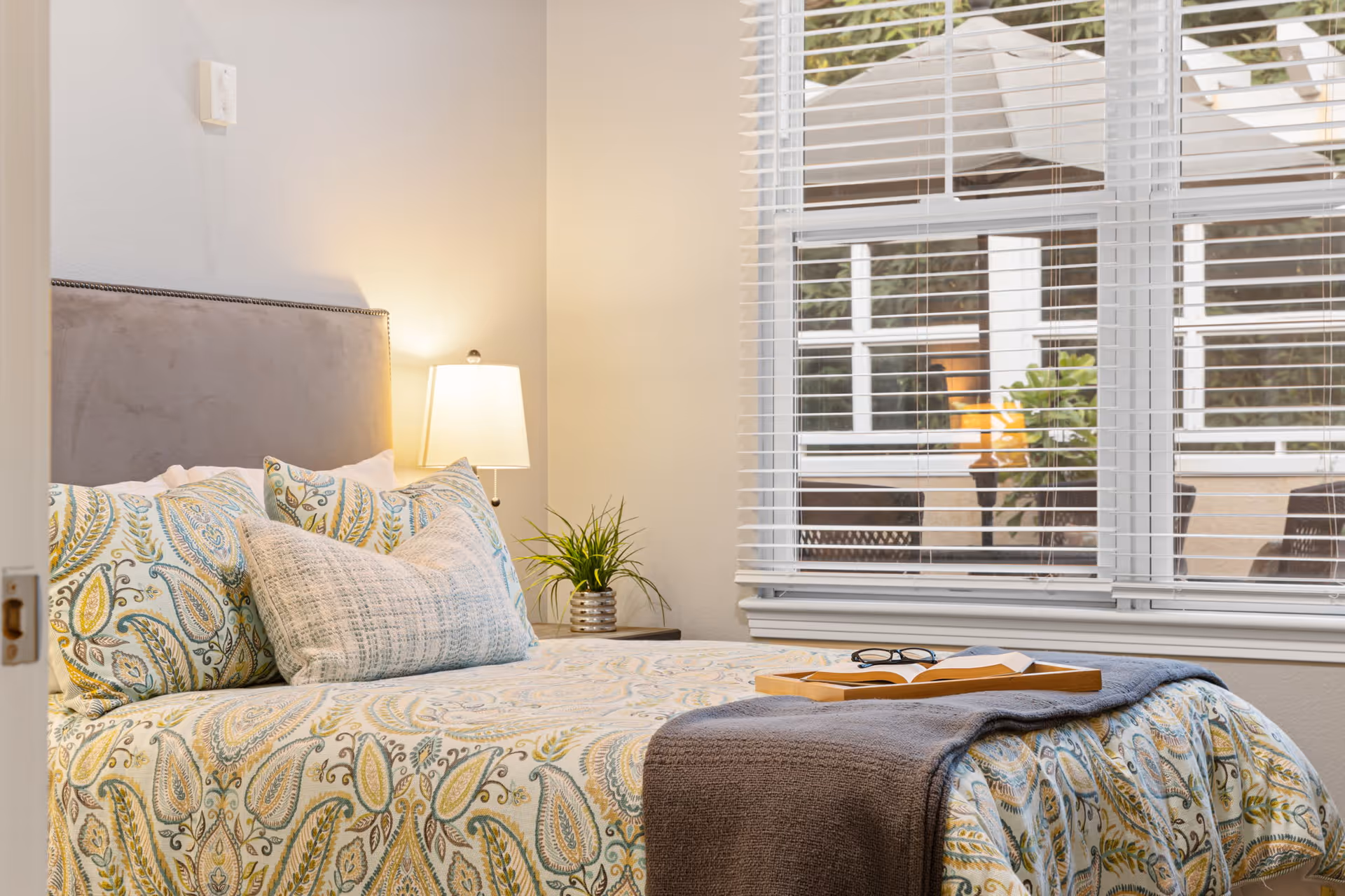 A cozy bedroom with a bed featuring a patterned bedspread and multiple pillows. A bedside table with a lamp and a small plant is next to the bed. A window with white blinds shows an outdoor patio area with a table and umbrella.