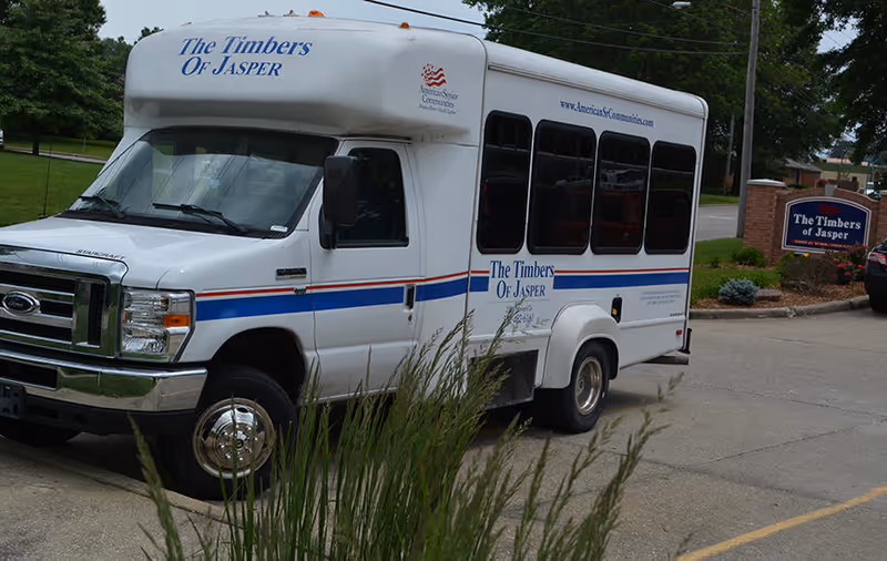 White shuttle bus labeled "The Timbers Of Jasper" parked outside the Timbers of Jasper facility with the facility sign visible.