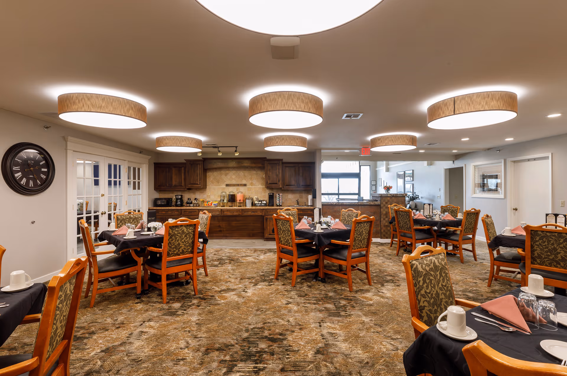 A dining room with multiple wooden tables and chairs arranged neatly. Each table is set with black tablecloths, pink folded napkins, white cups, plates, and silverware. The room has a patterned carpet and several large circular ceiling lights. At the back, there is a coffee bar area with wooden cabinets and various coffee and beverage supplies. A large wall clock is mounted on the left wall, and there are glass-paneled double doors on the left side.