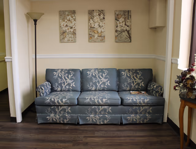 A living room area with a blue floral-patterned three-seater sofa against a beige wall. Above the sofa are three framed floral artworks. To the left of the sofa is a tall floor lamp, and to the right is a small wooden table with a floral arrangement. The floor is dark wood.