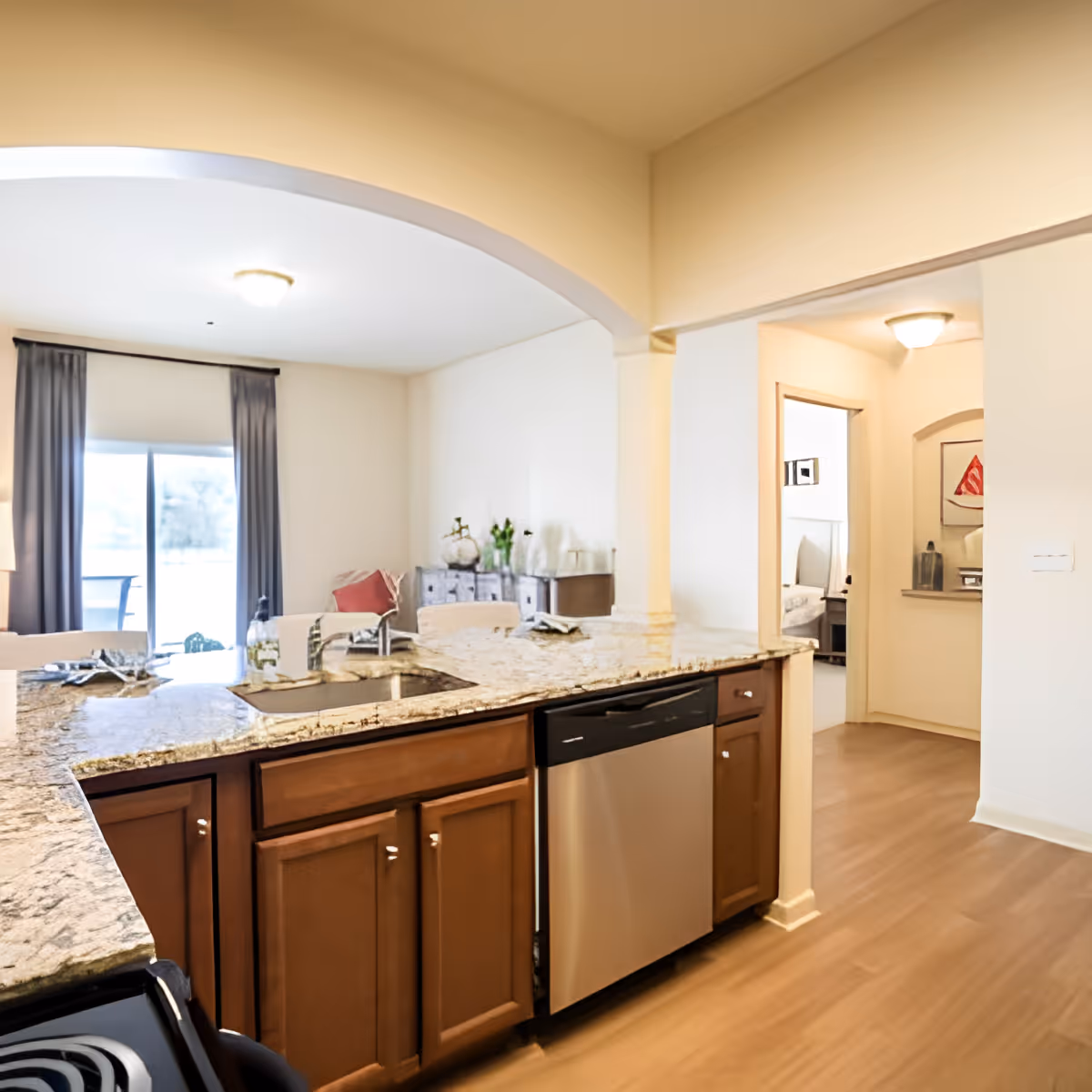 View of a modern kitchen with granite countertops, wooden cabinets, a stainless steel dishwasher, and a sink. The kitchen opens into a living area with a sliding glass door covered by dark curtains, and a glimpse of a bedroom is visible through an open doorway.