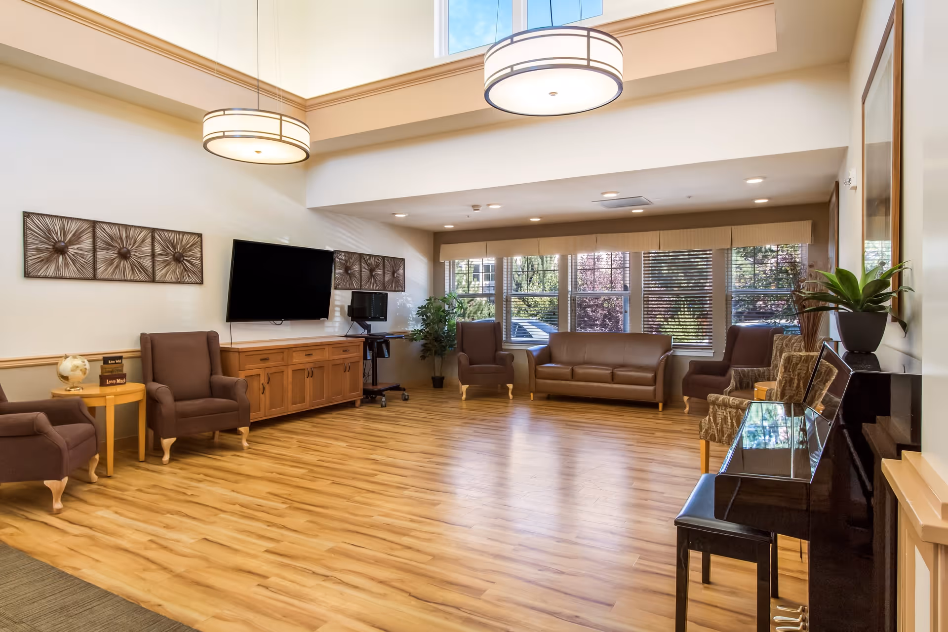 A spacious and well-lit living room area with wooden flooring, several brown armchairs and sofas arranged around the room, a flat-screen TV mounted on the wall above a wooden cabinet, large windows letting in natural light, two modern circular ceiling lights, decorative wall art, a small side table with a globe, and a black piano with a plant on top.