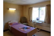 Simple single-occupancy patient bedroom with a hospital-style bed, armchair, window with curtains, and a wall light.