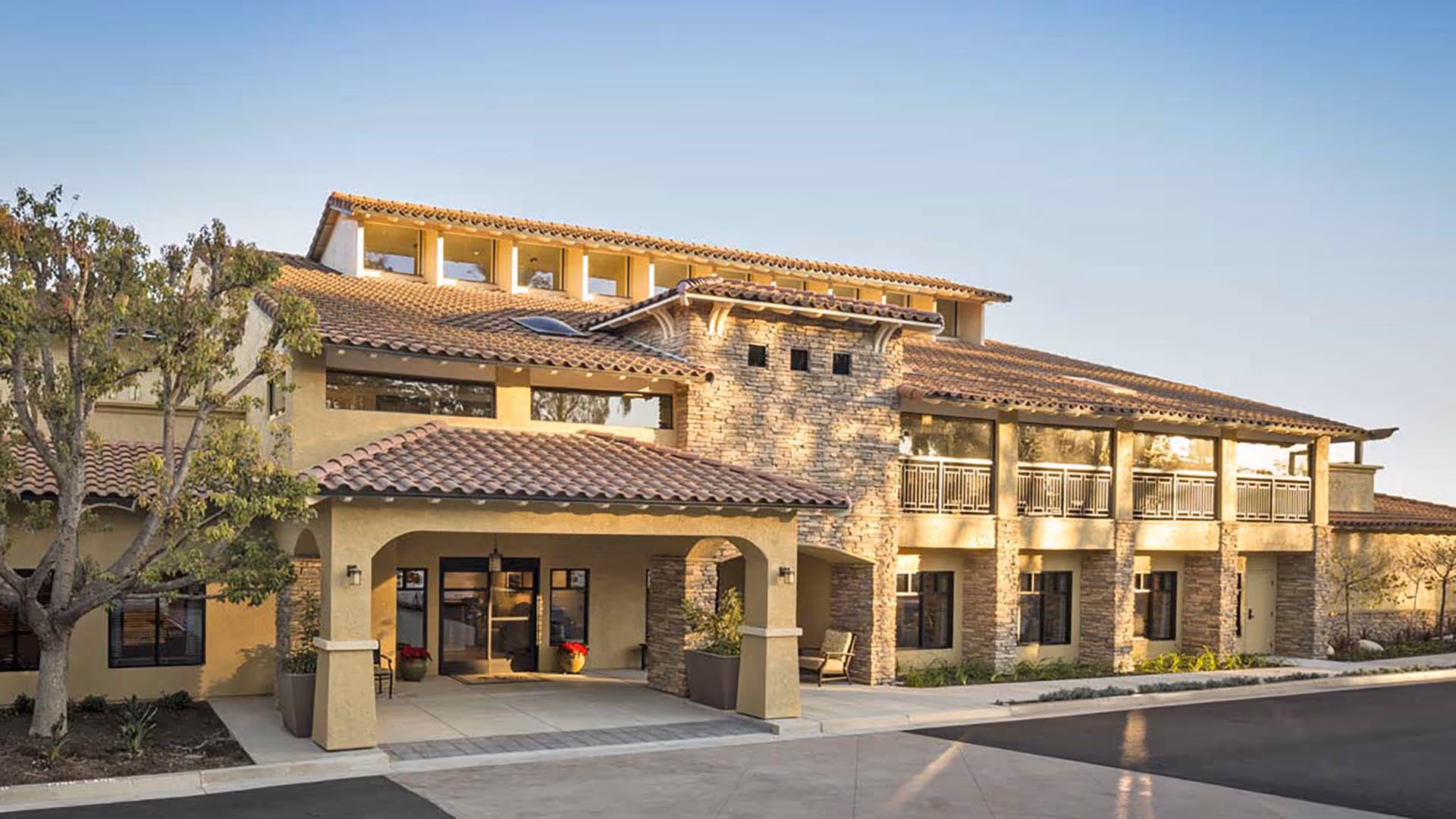Exterior view of a two-story senior living facility building with a tiled roof, stone and stucco walls, large windows, and a covered entrance. There are trees and plants around the building and a paved driveway in front.