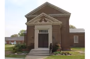 Front exterior view of a small brick building with a triangular pediment above the entrance, double doors with crosses on them, and steps leading up to the entrance. There is greenery and grass around the building.