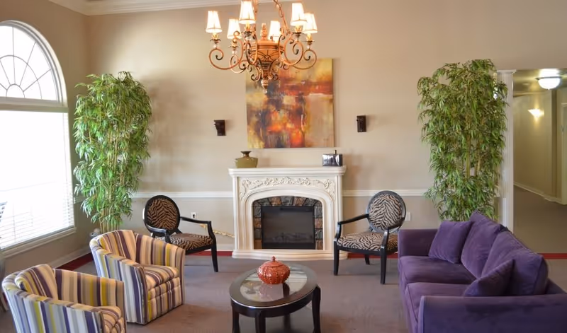 Cozy communal sitting area with a decorative fireplace, chandelier, purple sofa, patterned chairs, coffee table and tall potted plants.